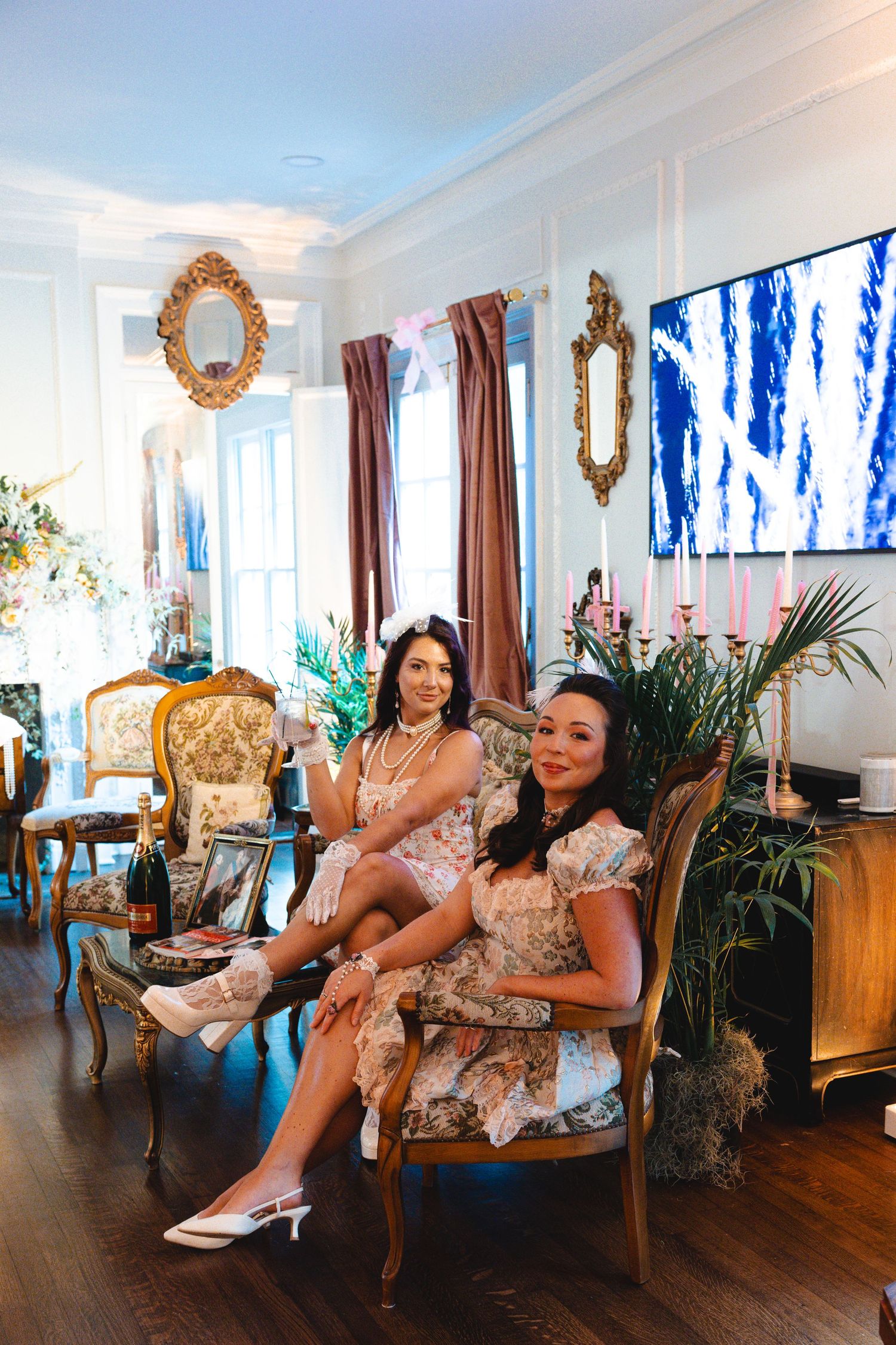Two elegant women in floral dresses pose in a luxurious French-style living room with gold mirrors.