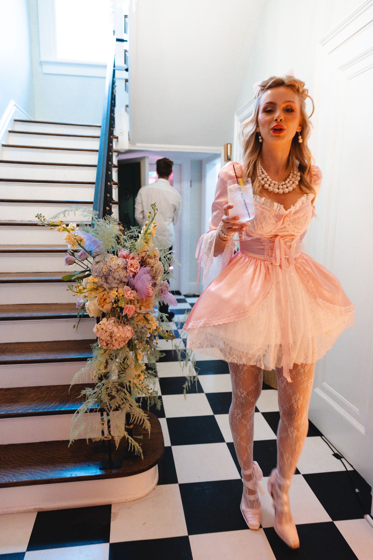 Blonde woman in pink mini dress holding champagne bottle poses by checkered floor staircase with pastel floral arrangement.
