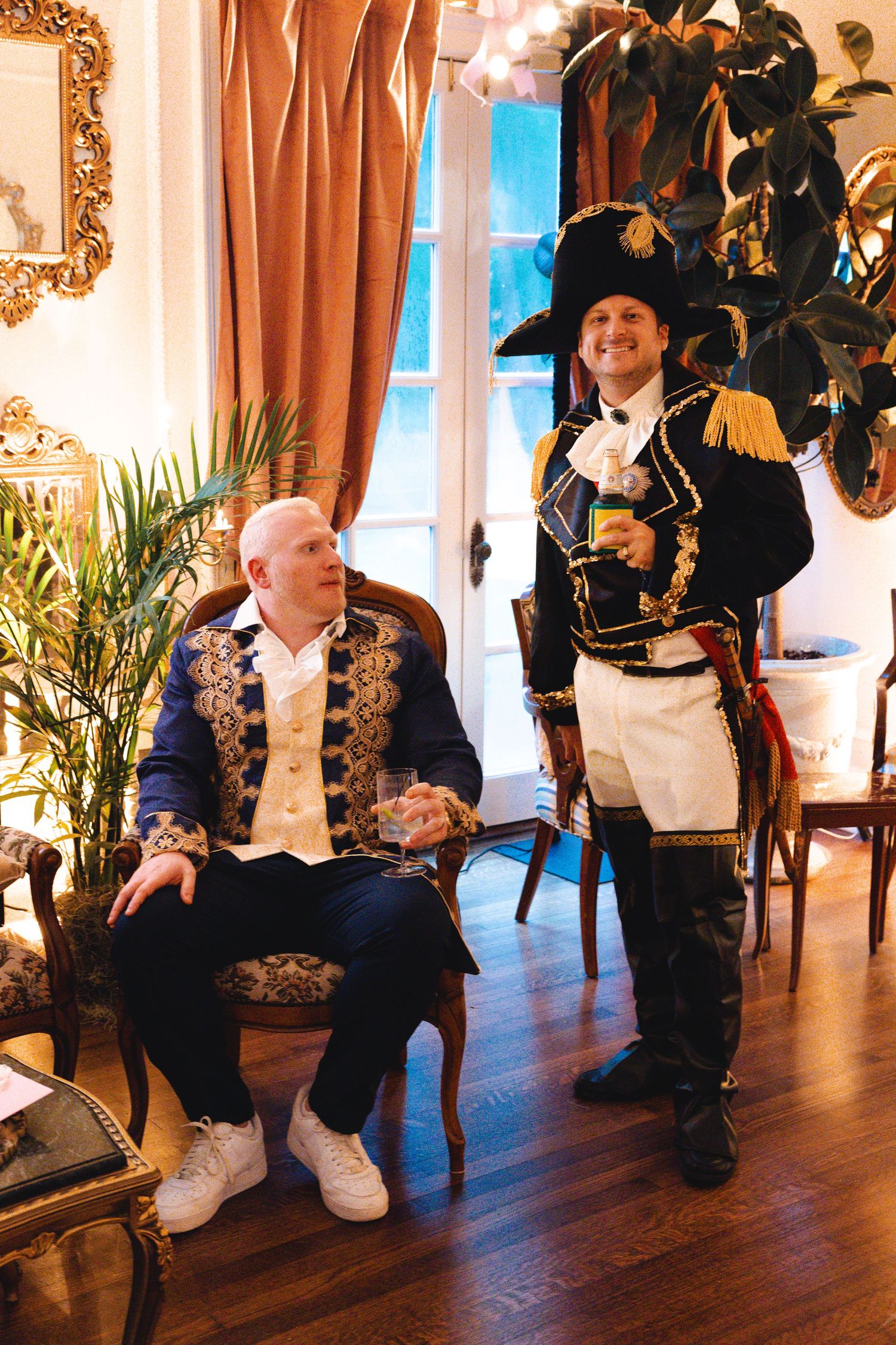 Two men in ornate historical costumes pose in an elegant room with gold-framed mirrors and drapes.