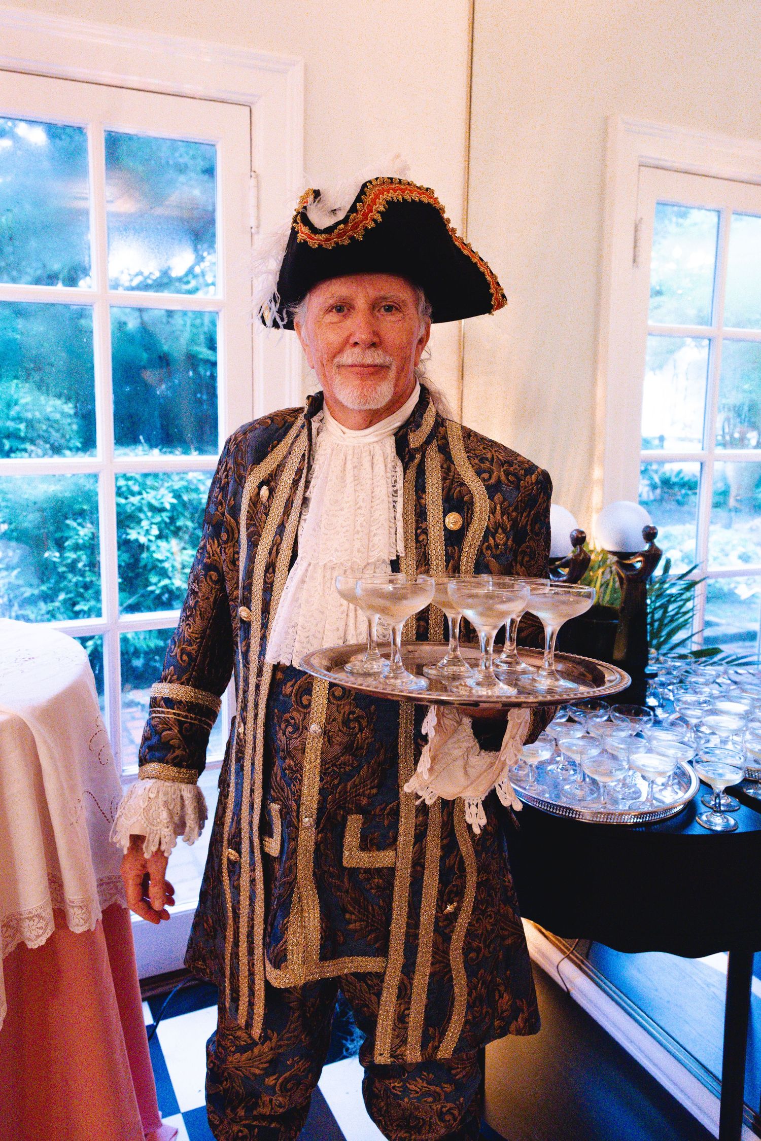 An elderly man in an ornate colonial costume and tricorn hat holds a tray of champagne glasses at a formal event.