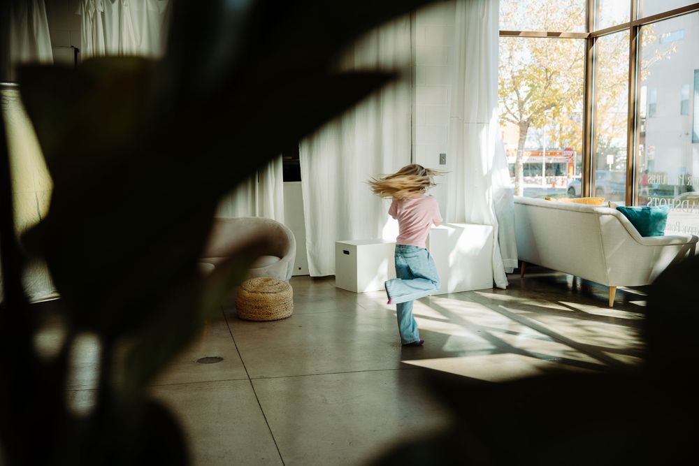 Young girl dancing in sunlit living room with large windows and white curtains on a hardwood floor.
