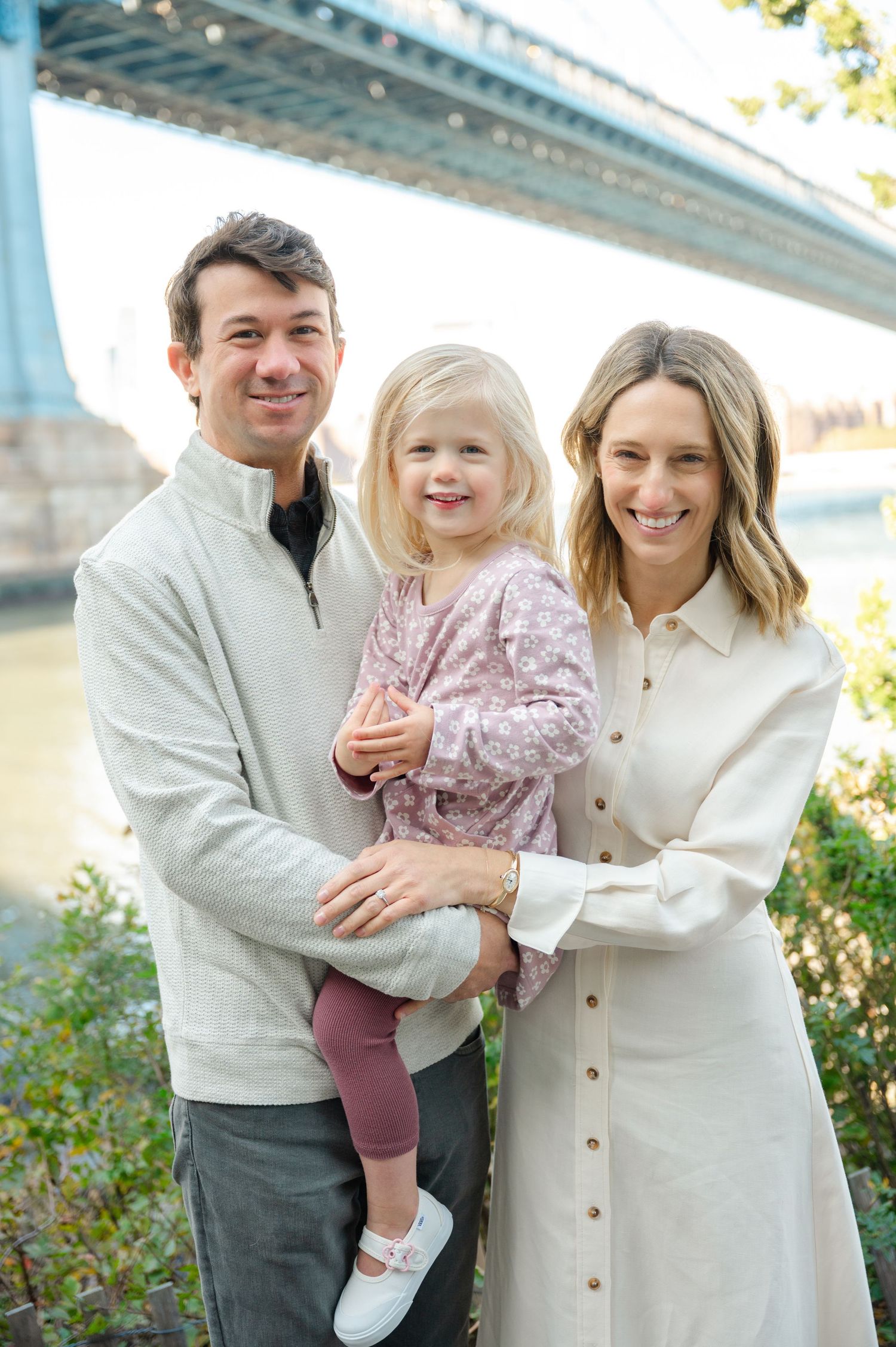A smiling family of three poses near a bridge, parents holding their young blonde daughter outdoors.