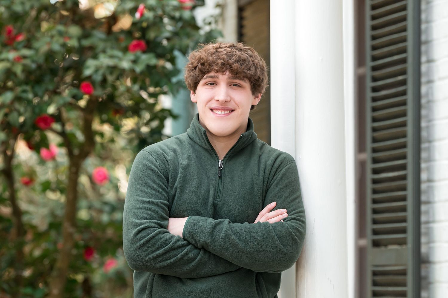 Smiling young man with curly hair in green zip-up standing with arms crossed near red flowers.