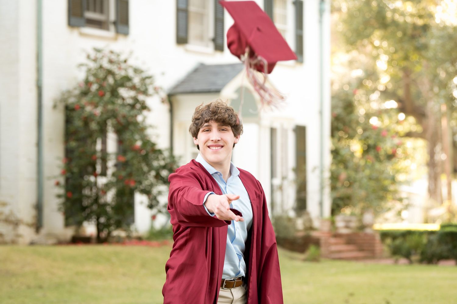 Smiling young man in red graduation gown tossing his cap in front of a white historic building.