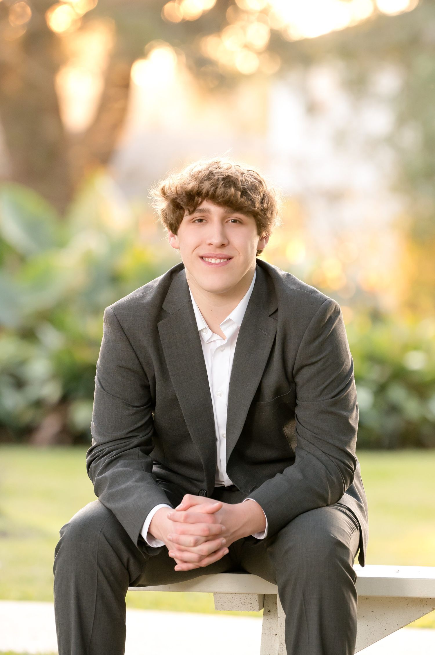 Smiling young man in dark suit sitting on bench with bokeh garden background.