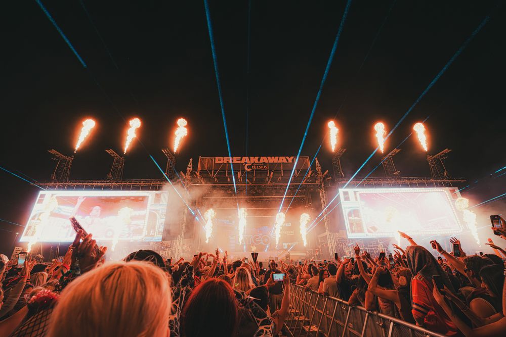 Crowd at Breakaway Music Festival enjoying a night concert with dramatic light beams and fire effects on stage.