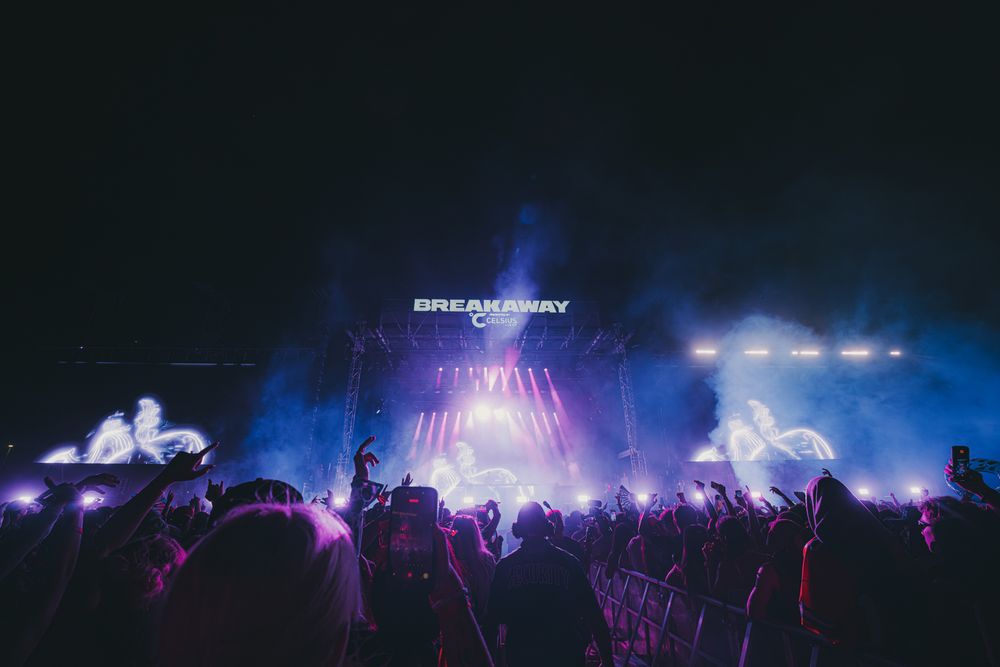 Breakaway music festival crowd at night with purple lights and illuminated stage.