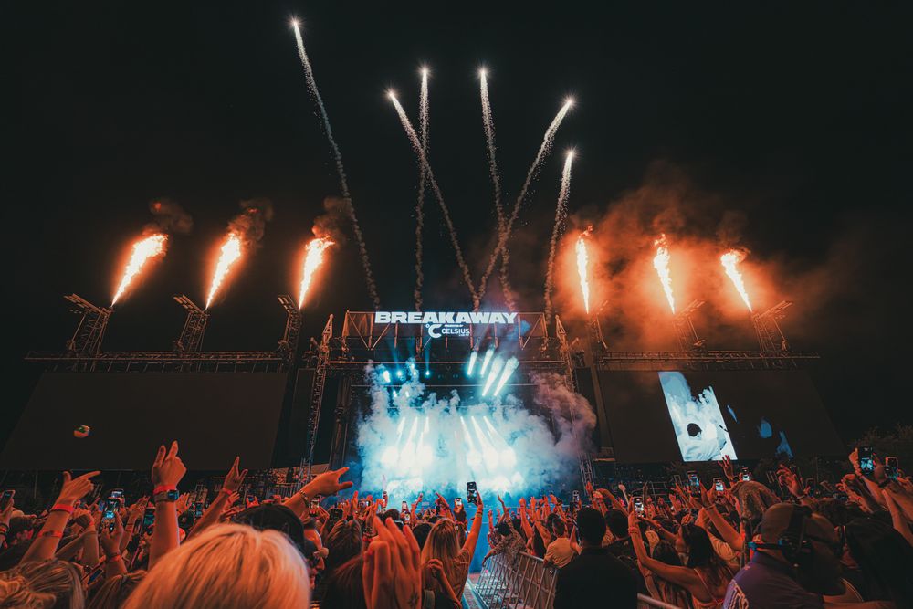 Crowd cheering at outdoor music festival with fireworks and stage lights illuminating the night sky.