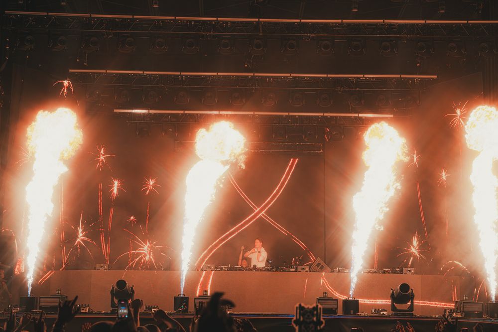 DJ performs on stage with massive pyrotechnic fire blasts and fireworks at a large outdoor music festival.