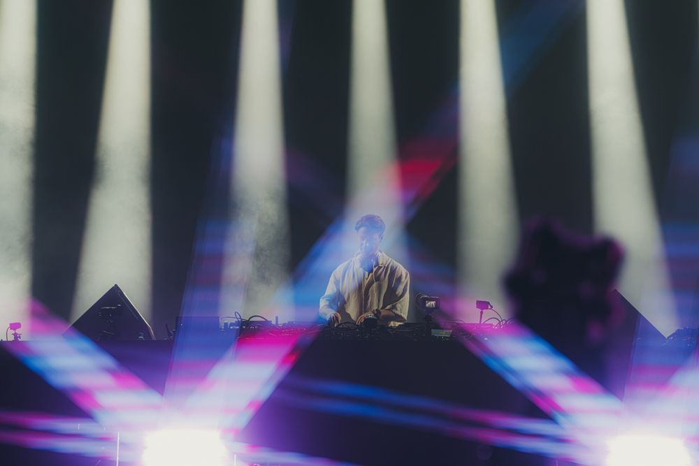 DJ performing on stage surrounded by dramatic beams of purple and white light at a live music concert.