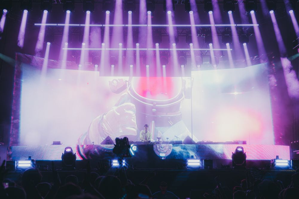 A DJ performs on a massive stage with dramatic purple and white light beams at a packed concert venue.