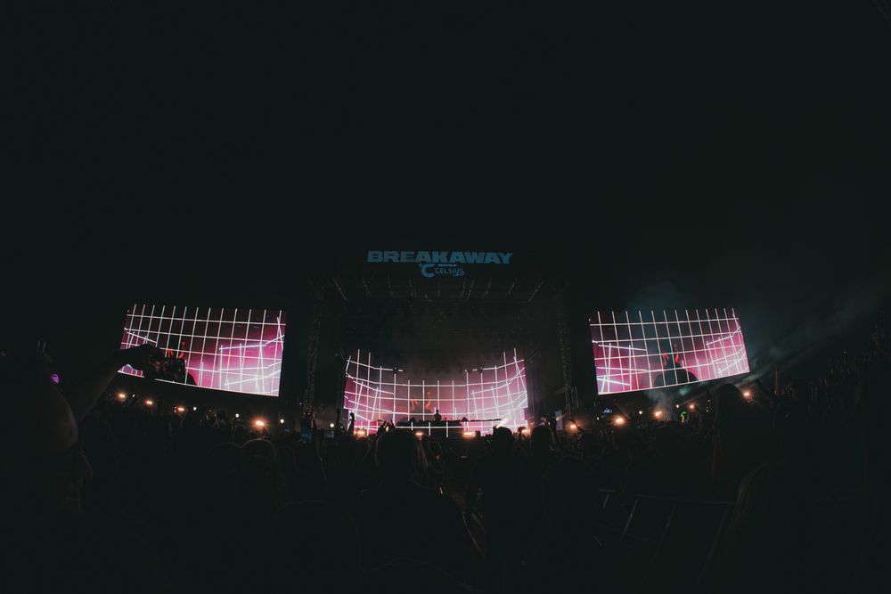 Concert stage with large LED screens displaying pink and white visuals, surrounded by a dark crowd at a live music event.