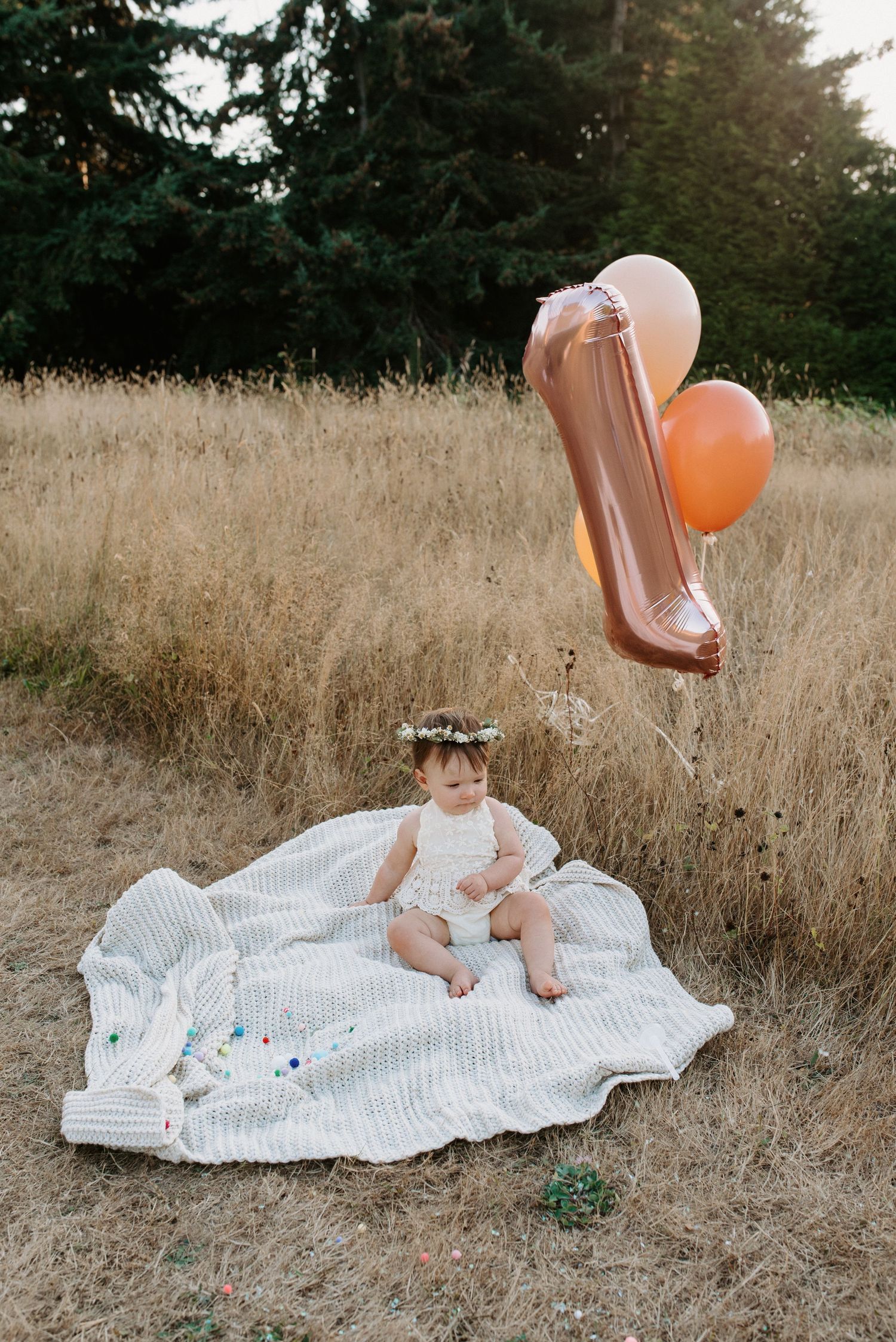 Baby girl in white romper and flower crown sits on blanket in field with number one balloon for first birthday.