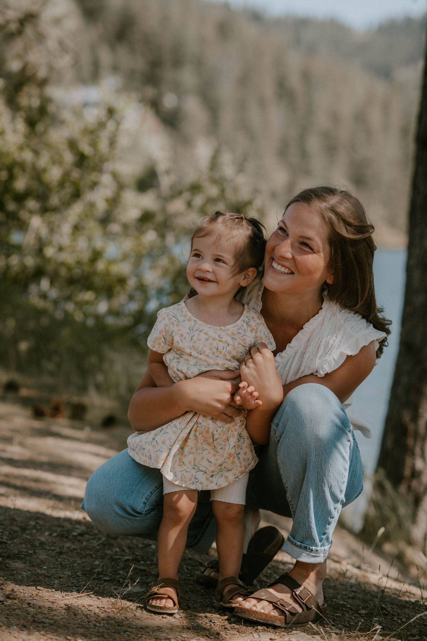 A smiling mother crouches outdoors holding her happy toddler daughter, both wearing casual outfits near a scenic lake.