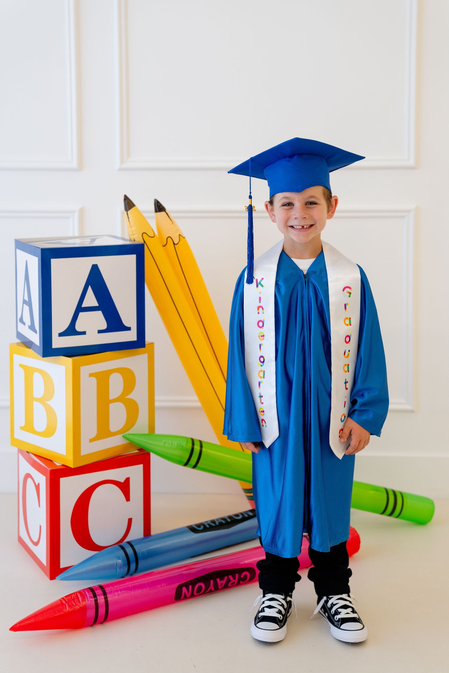 Young boy in blue kindergarten graduation cap and gown poses with ABC blocks and giant crayons and pencils.