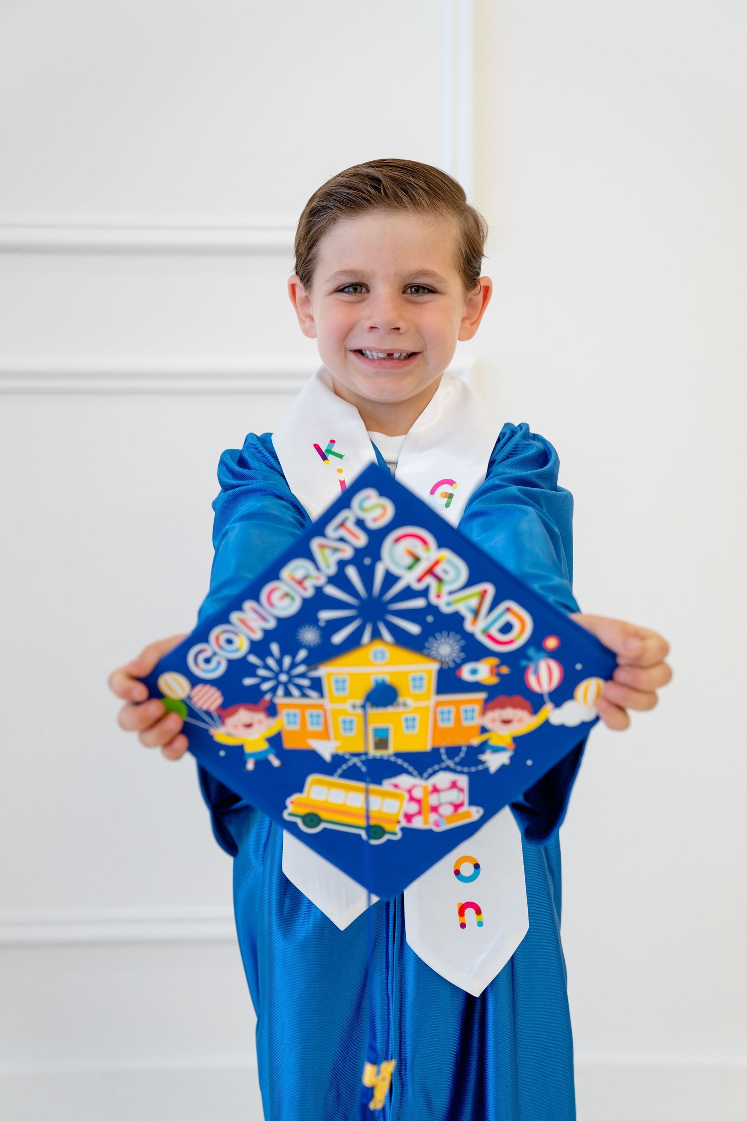 Smiling young boy in blue graduation gown holding a decorative Congrats Grad cap with school-themed artwork.