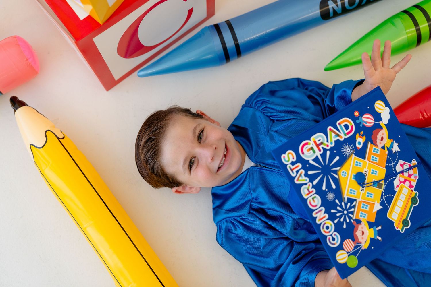 A smiling boy in a blue graduation hoodie lies surrounded by oversized colorful crayons and a pencil on a white background.