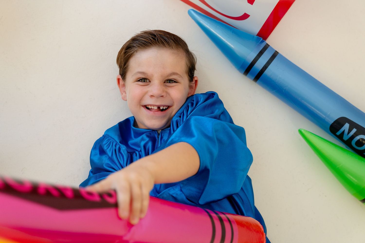 Smiling boy in blue raincoat lying among oversized colorful crayon props for a fun back-to-school photo.