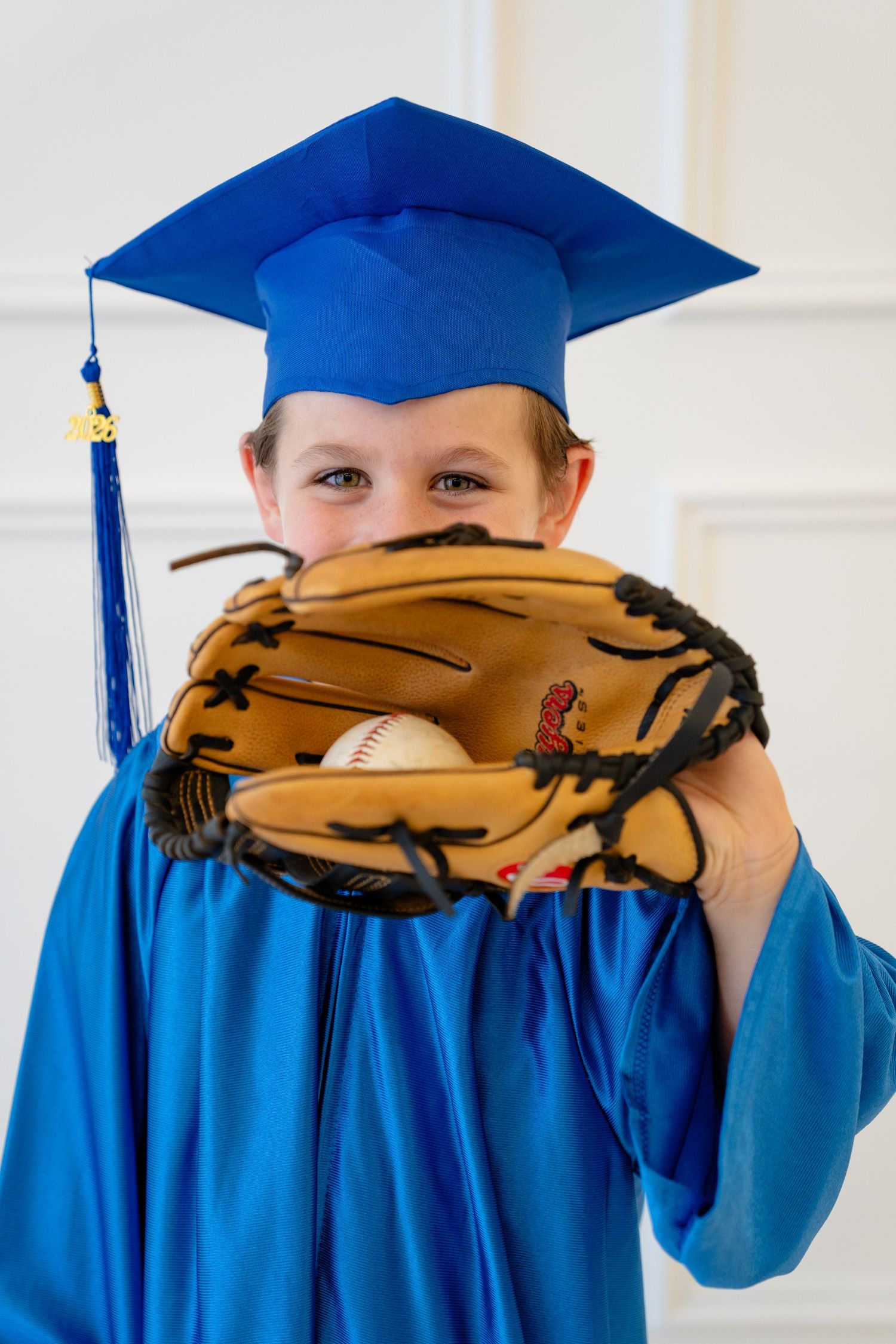 Young child in blue graduation cap and gown holding a baseball glove with ball, smiling at camera.