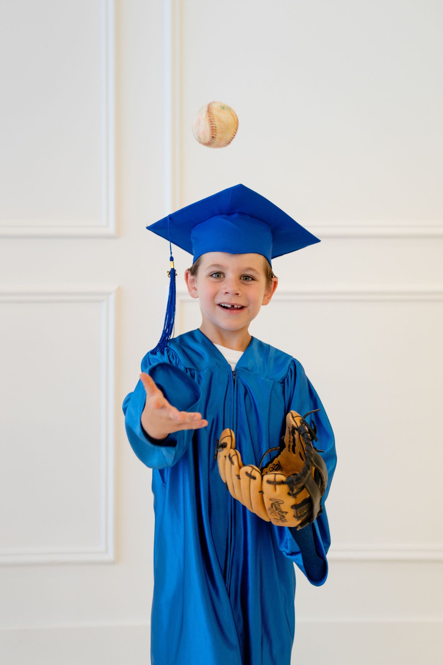 A smiling boy in a blue graduation cap and gown tosses a baseball while wearing a leather mitt.