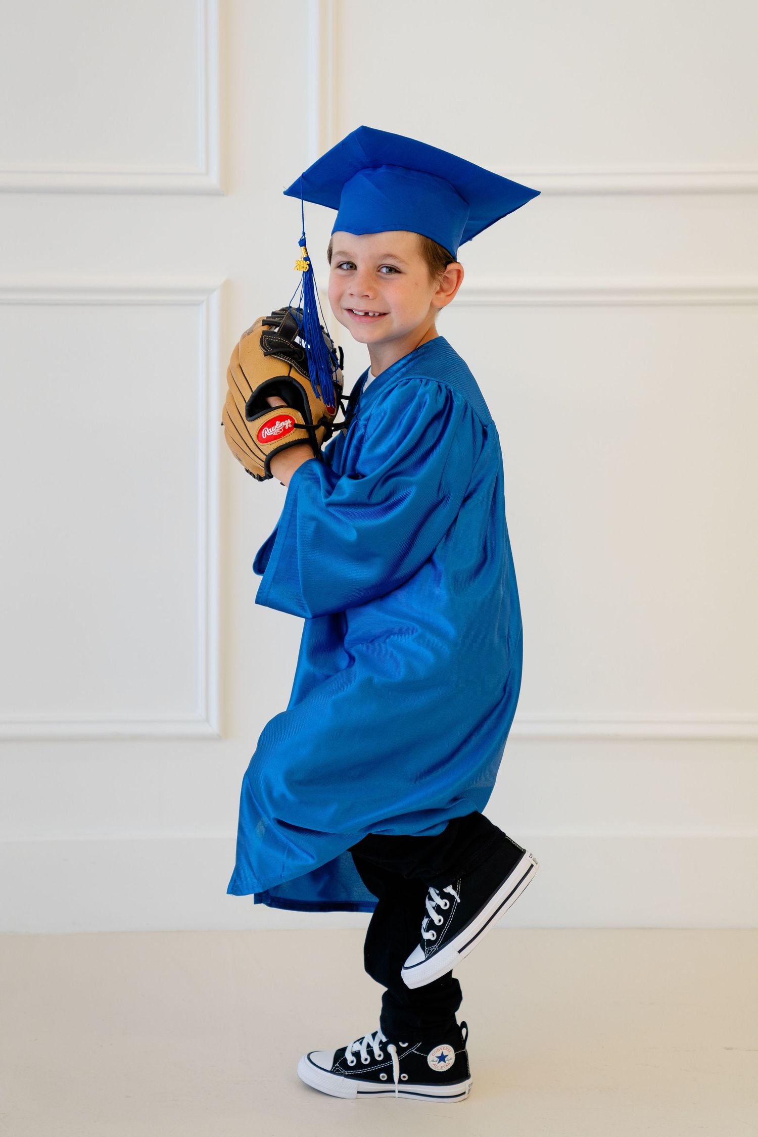 Young boy in blue graduation cap and gown posing with a baseball glove wearing black Converse sneakers.