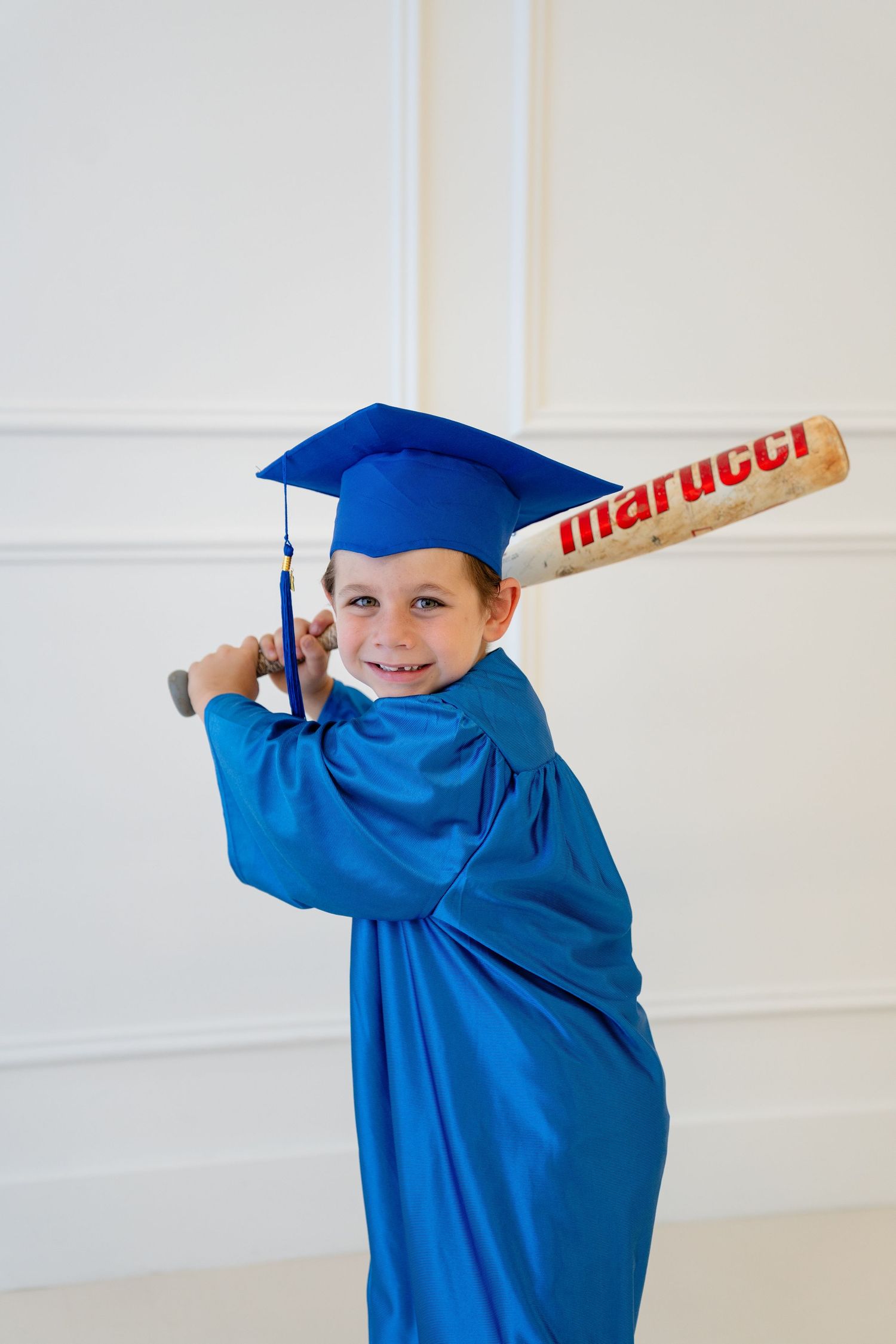 Young boy in blue graduation cap and gown smiling while holding a Marucci baseball bat in batting stance.
