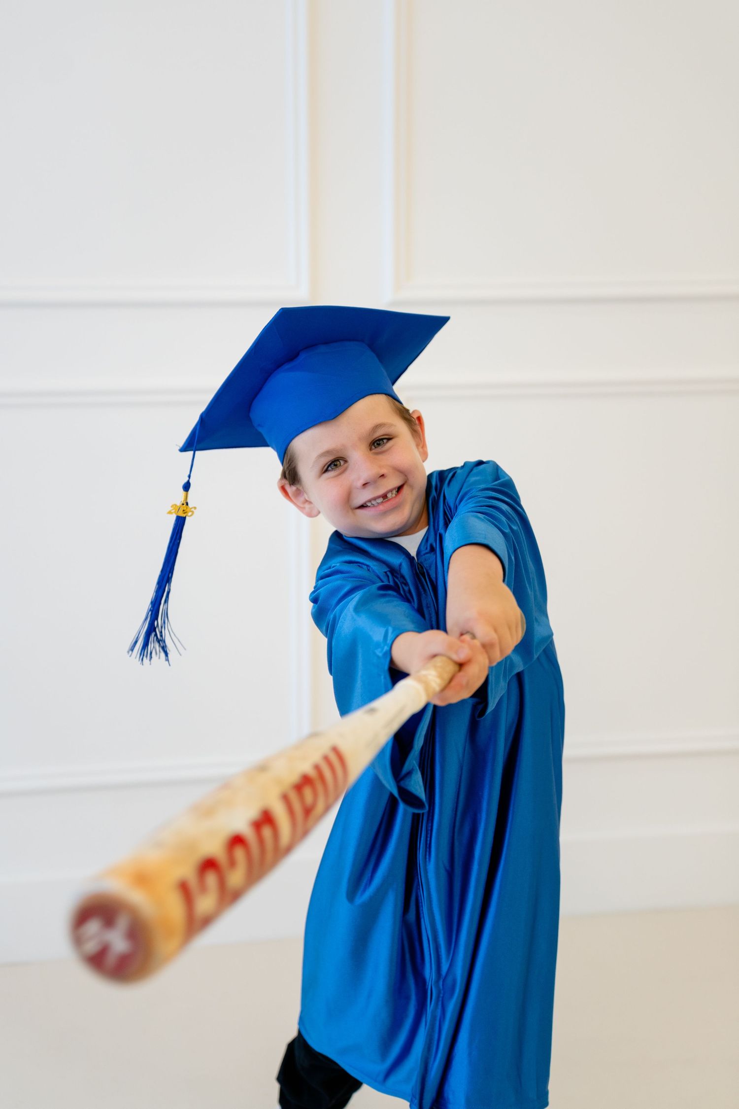Smiling child in blue graduation cap and gown swinging a wooden baseball bat indoors.