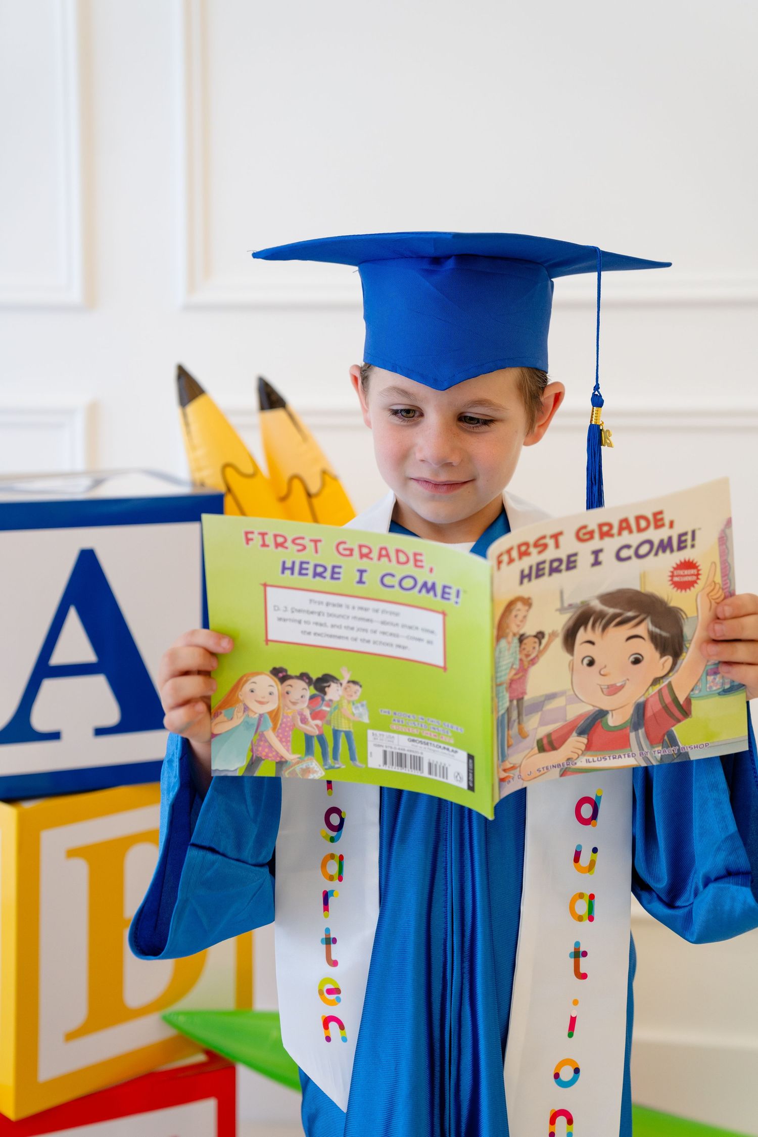 Child in blue graduation cap and gown reading First Grade Here I Come book, surrounded by colorful alphabet blocks.