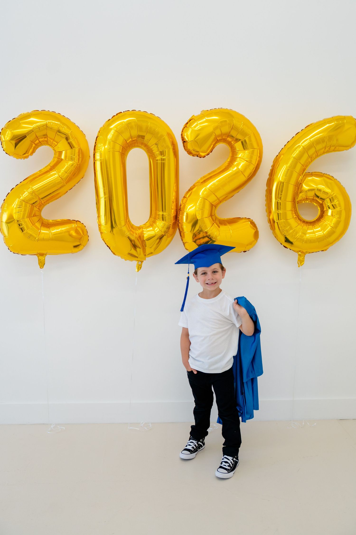 Young boy in blue graduation cap holding gown, posing in front of gold 2026 balloon numbers on white wall.