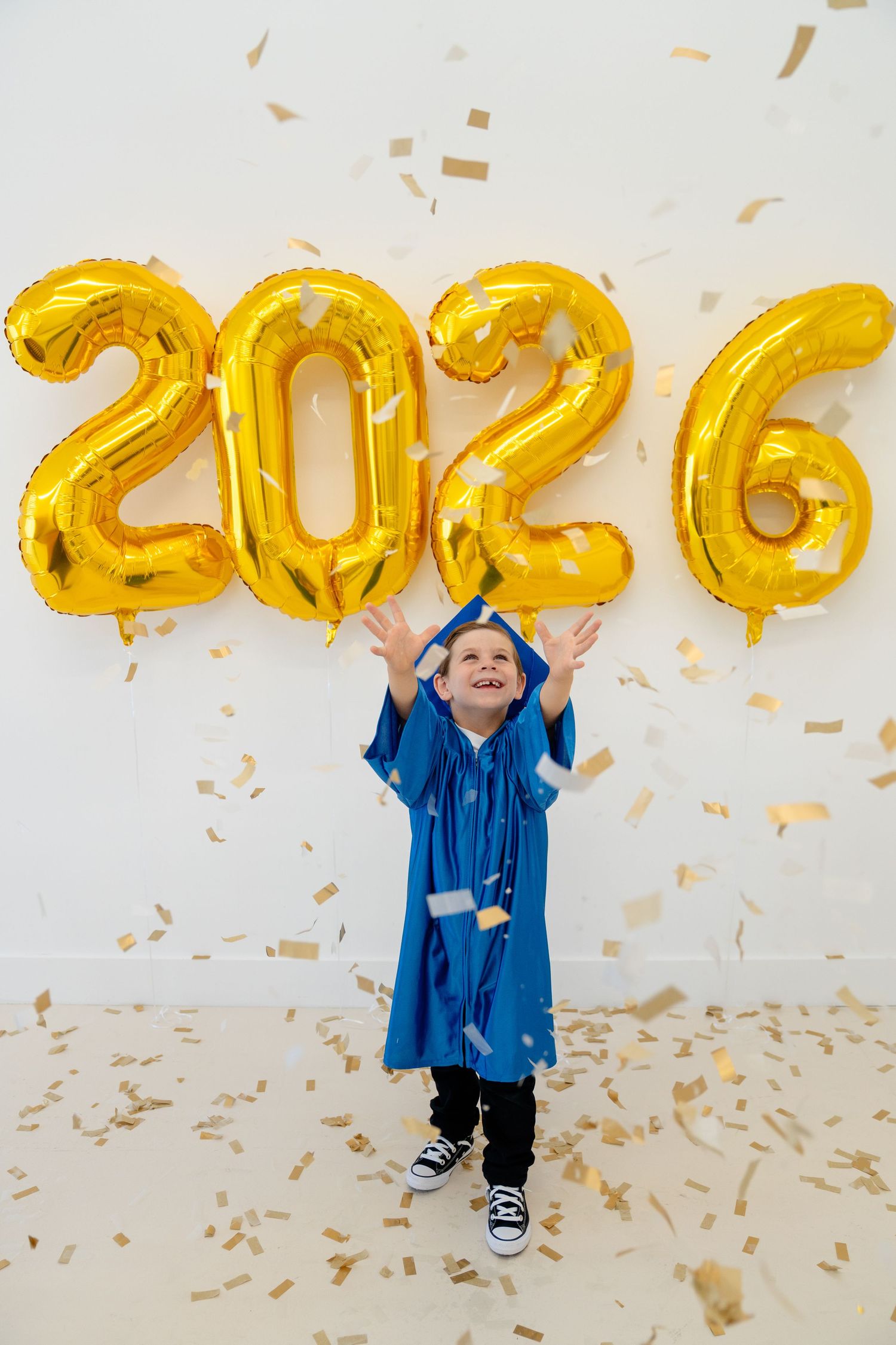 Young boy in blue graduation gown celebrates with gold 2026 balloons and falling confetti.
