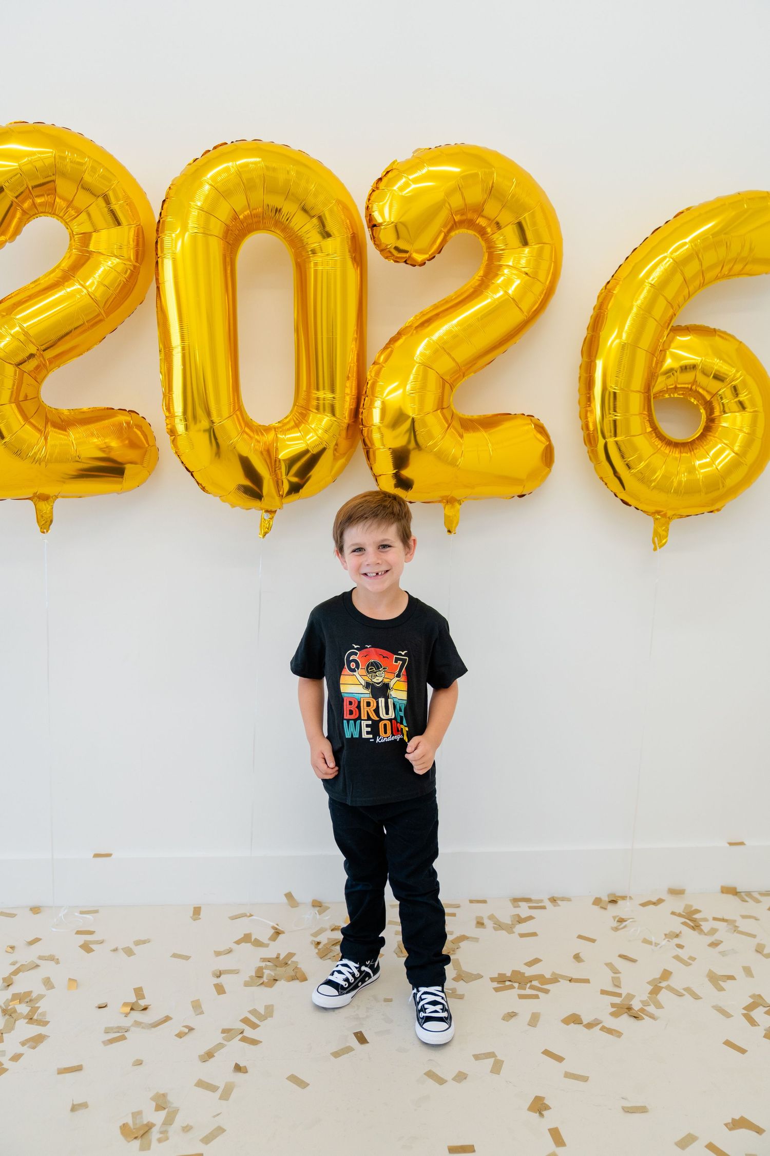 Young boy smiling in front of gold 2026 balloon numbers with confetti on floor wearing black graphic tee.