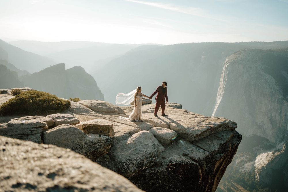 Couple in wedding attire holding hands on a rocky cliff at Yosemite with stunning mountain valley views.