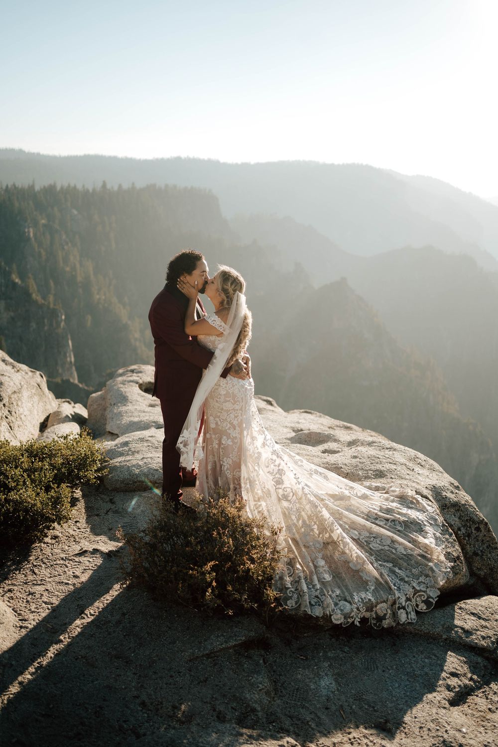 Bride and groom share a kiss on a rocky cliff at sunset with misty mountain forest views in Yosemite.