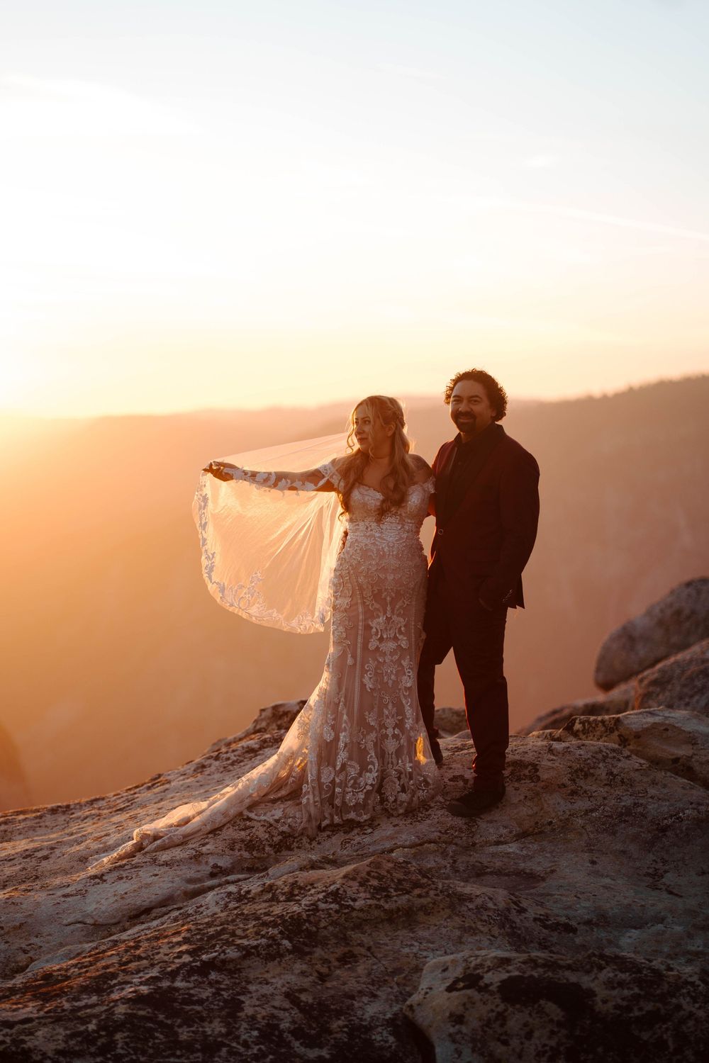 Bride and groom stand on mountain cliff at golden sunset, her lace gown and veil flowing in the wind.