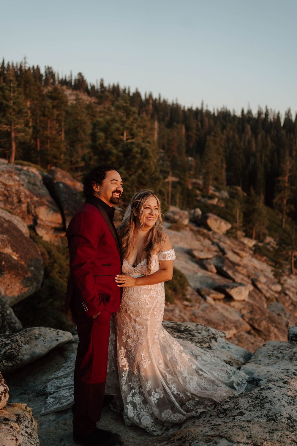 A joyful couple in wedding attire poses on rocky terrain with pine trees at golden hour.