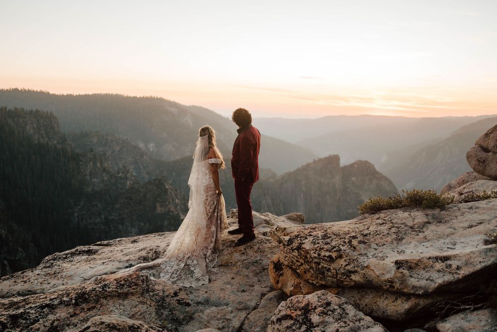 Bride and groom stand on rocky cliff at sunset overlooking Yosemite Valley in breathtaking outdoor wedding ceremony.