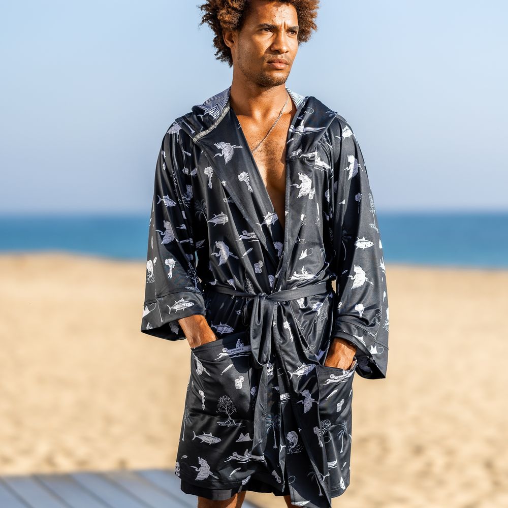 Man wearing a dark navy shark-print belted robe on a sunny beach with ocean in the background.