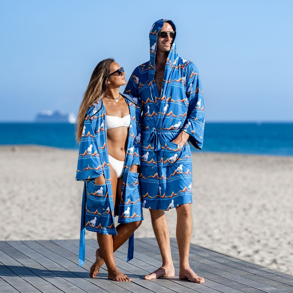 Couple wearing matching blue printed hooded robes on beach boardwalk, woman in white bikini underneath.