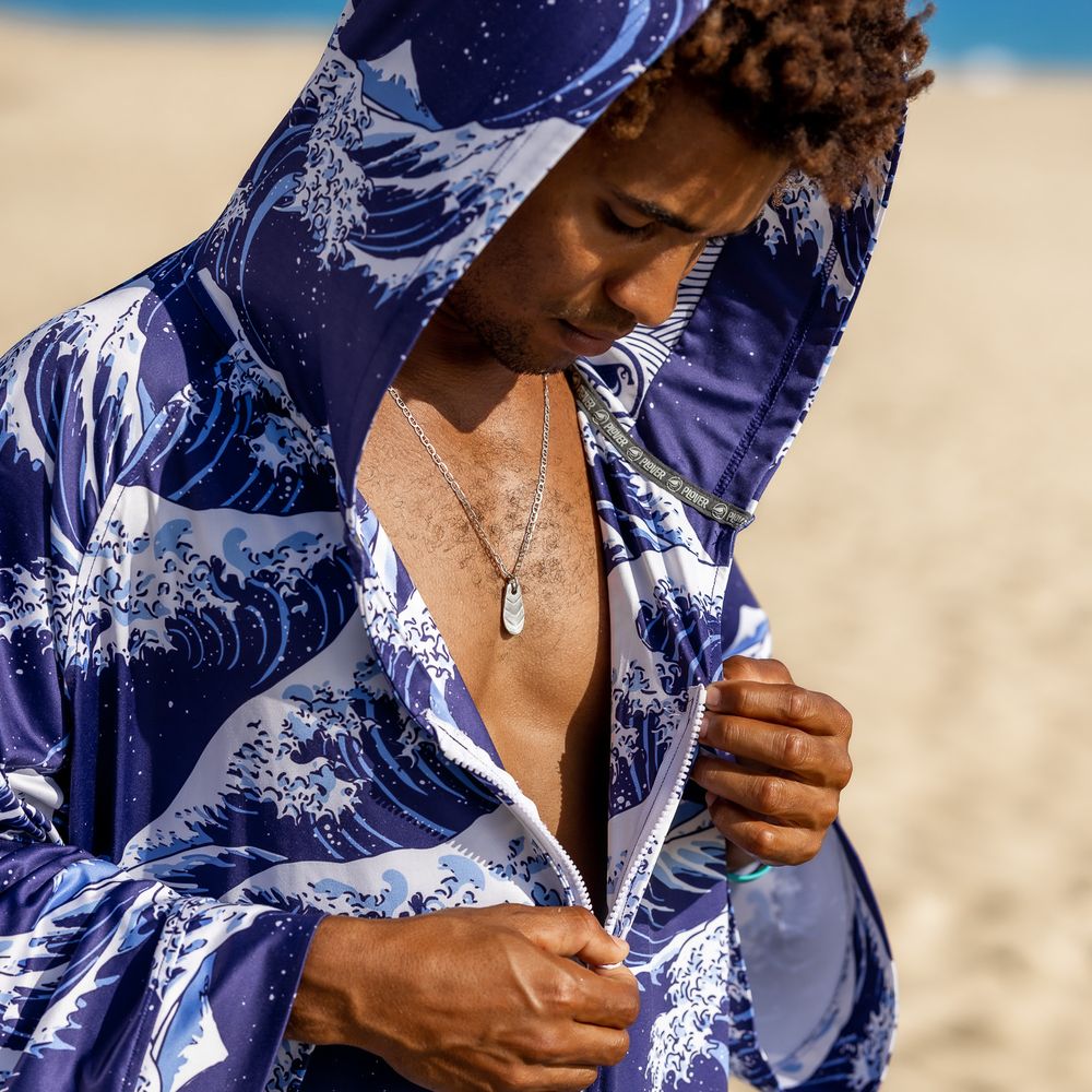 Man wearing a blue and white wave-print hooded jacket on a sunny beach, showcasing a nautical necklace.