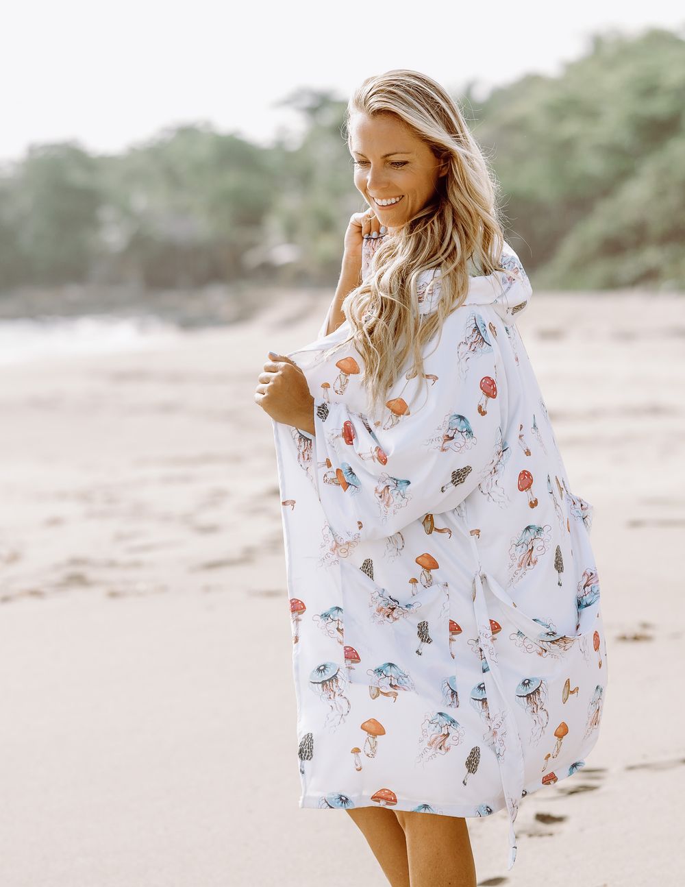 Woman on beach wearing white jellyfish print kimono cover-up, smiling and holding fabric edges.