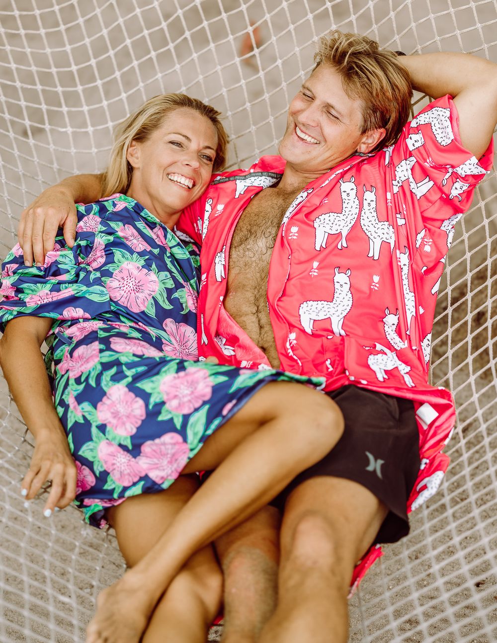 Couple laughing and relaxing together in a hammock wearing colorful printed robes outdoors.