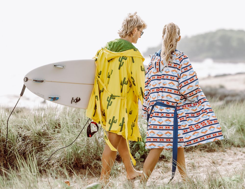 Two surfers wearing colorful hooded ponchos walk on a sandy beach, one carrying a white surfboard.