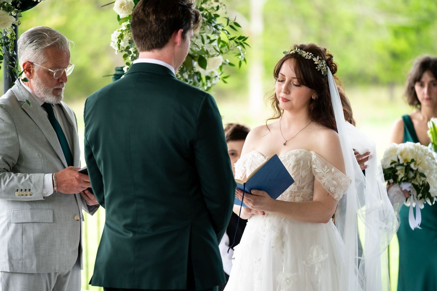 Bride and groom exchange vows outdoors with officiant, bride wearing lace gown and floral crown at elegant wedding ceremony.