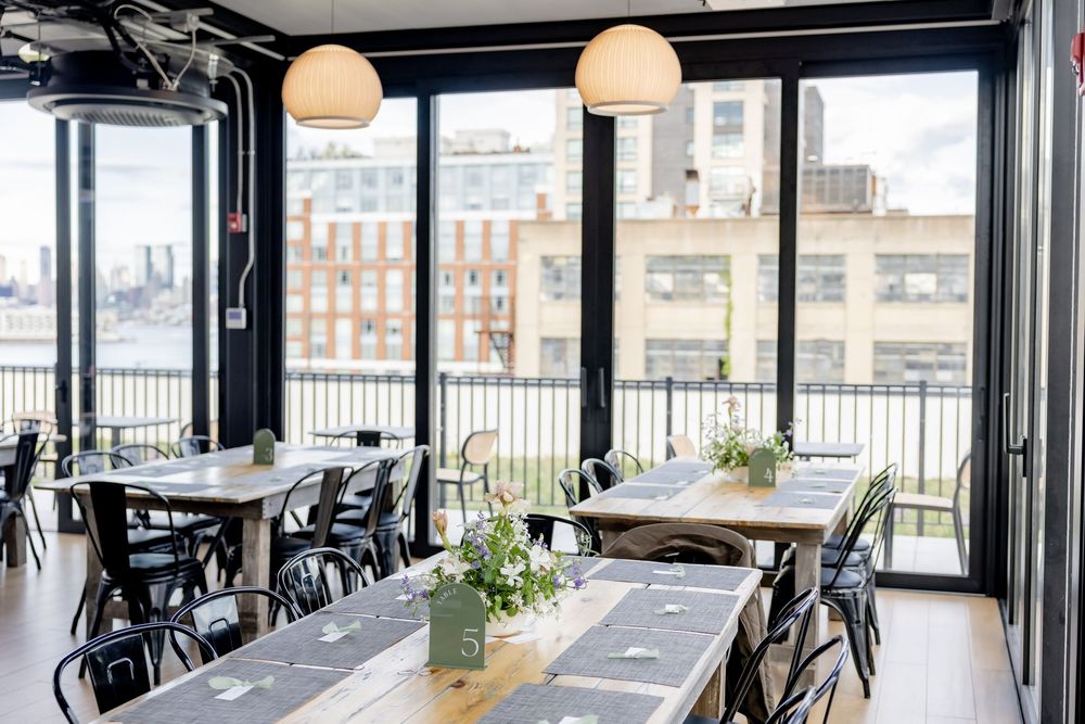 Bright rooftop restaurant dining room with floor-to-ceiling windows, black chairs, wooden tables, and city skyline views.