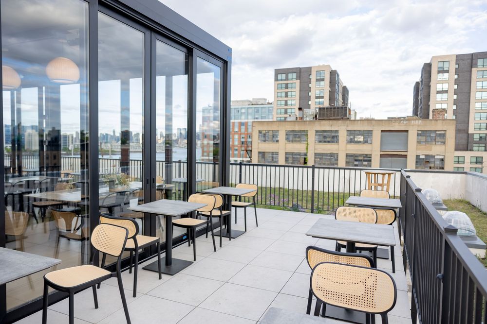 Modern rooftop patio with black metal chairs, small tables, glass walls, and urban skyline views on a clear day.