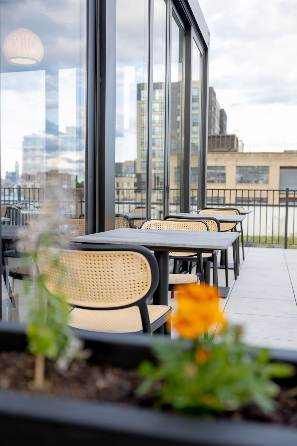 Rooftop patio dining area with rattan chairs, dark tables, glass walls, and city skyline views on a cloudy day.