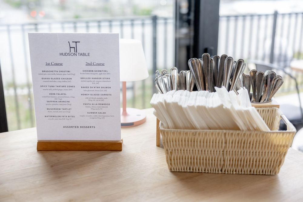 Hudson Table restaurant menu card beside a wicker basket of cutlery and napkins on a wooden table.
