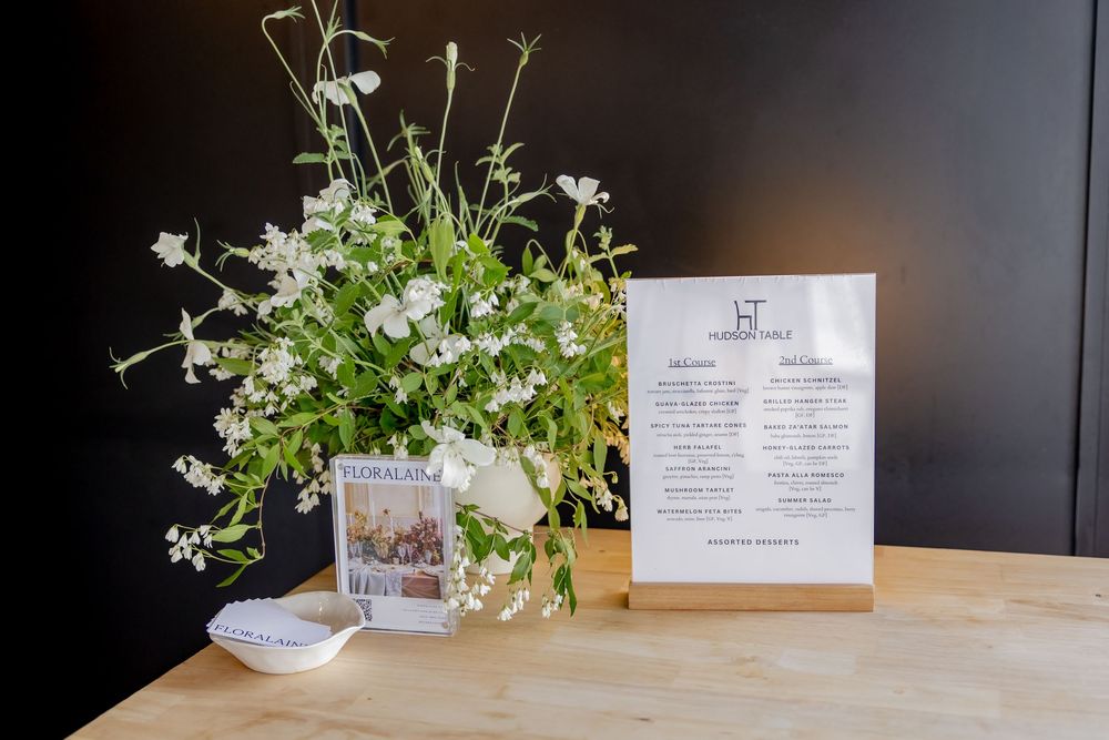 White wildflowers in a glass vase beside a menu card and magazine on a wooden table against a dark wall.