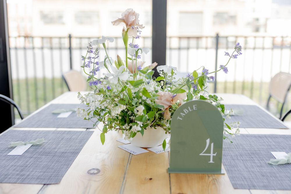 Wedding table 4 centerpiece with white bowl of pink roses, purple wildflowers, and greenery on rustic wood table.