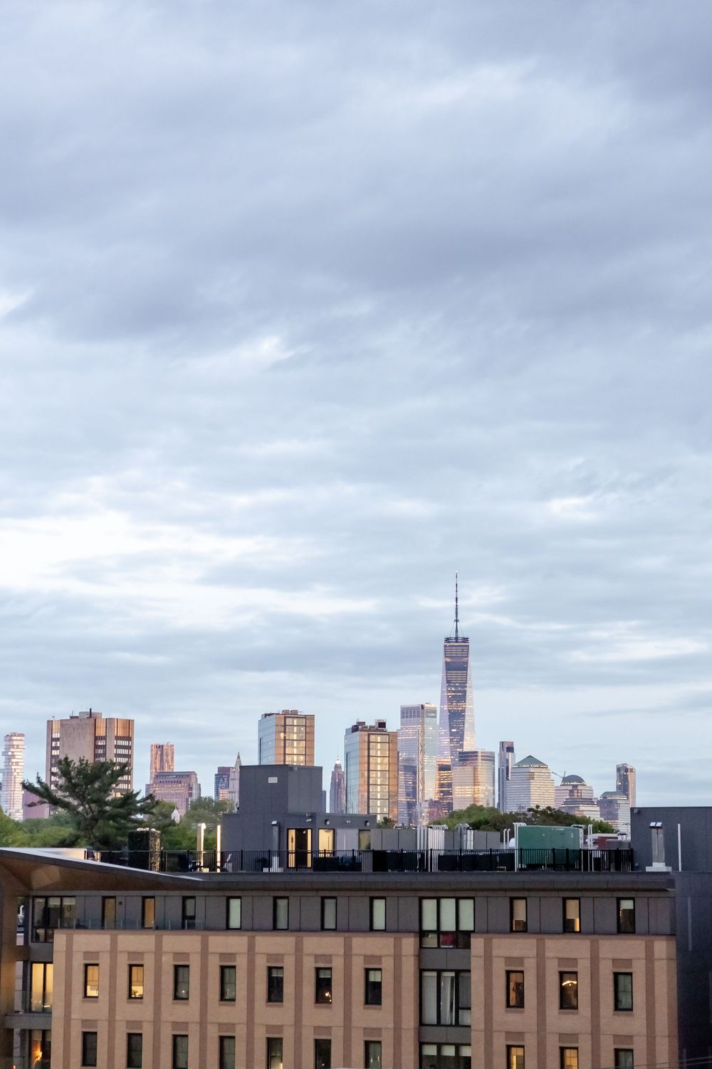 NYC skyline with One World Trade Center visible behind foreground buildings under cloudy sky at dusk.